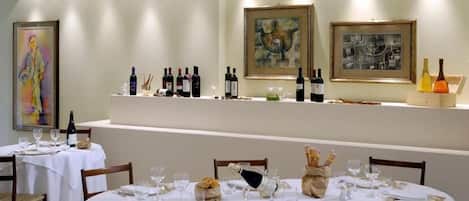 Un comedor con mesas redondas dispuestas para una comida, con manteles blancos, cubiertos ordenados y botellas de vino en una estantería.