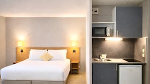Ein Hotelzimmer mit Bett, Nachttischlampen, einem kleinen Holznachttisch, einer integrierten Küche inklusive Mikrowelle, Spüle und Kühlschrank.