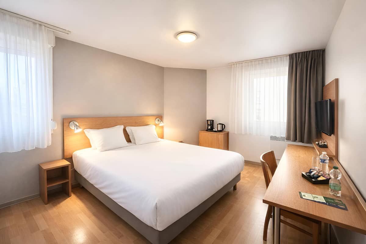 Ein Hotelzimmer mit einem großen Bett, Nachttischen, einem Schreibtisch mit Stuhl, einem Flachbildfernseher und einem Fenster mit Vorhängen.