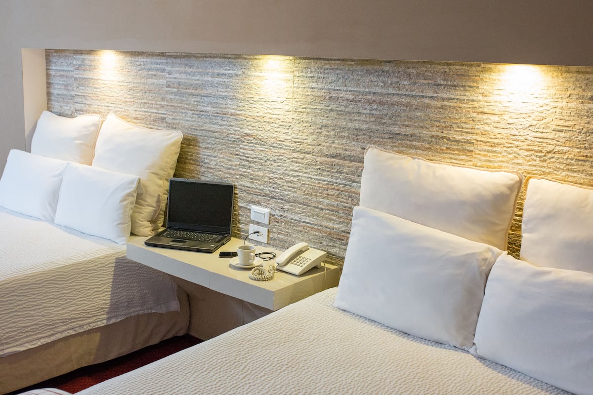 A hotel room with two beds, a desk with a laptop, a phone, and a cup.