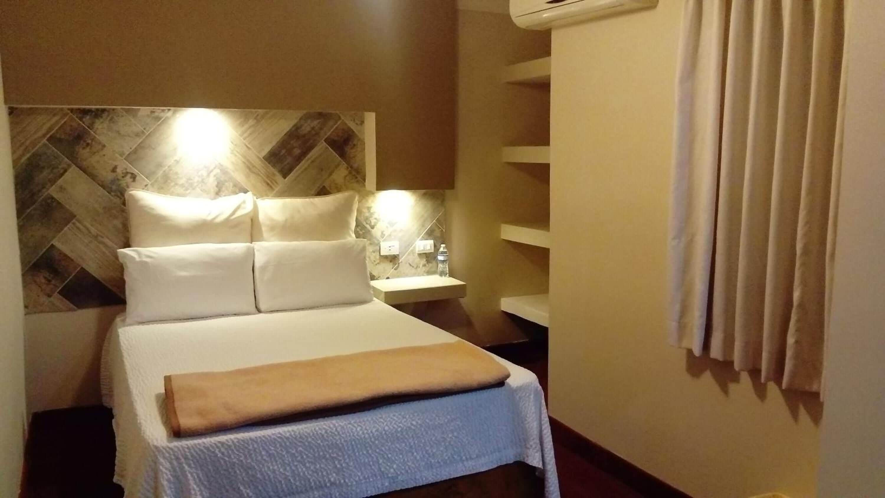 A hotel room with a bed, a nightstand, a wall-mounted shelf, and a curtain.