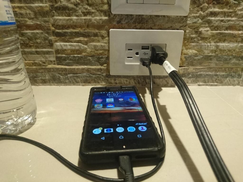 Un teléfono inteligente cargándose sobre una superficie blanca, con una botella de agua cerca.