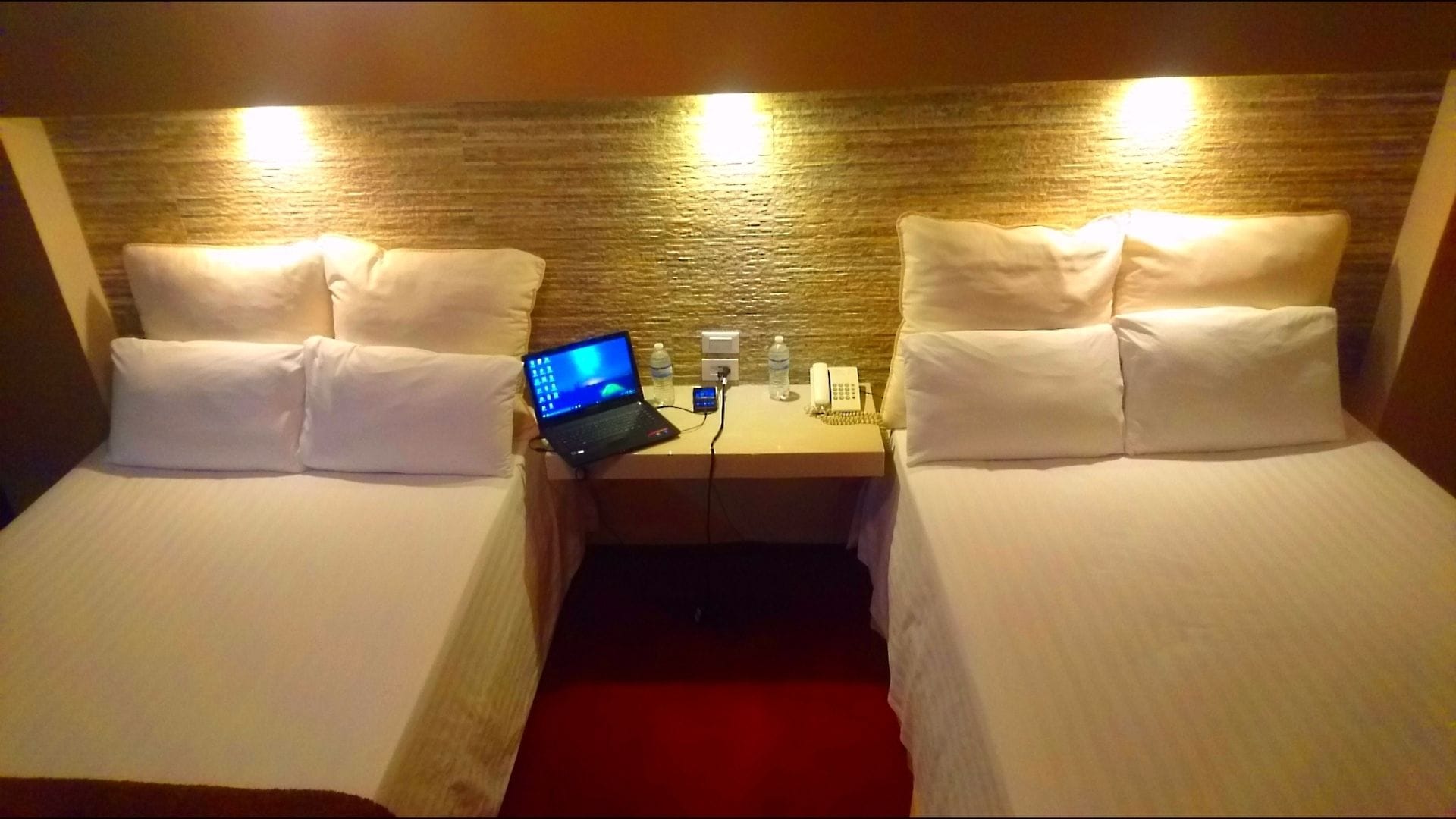 A hotel room with two single beds, a desk with a laptop, a phone, and a water bottle.
