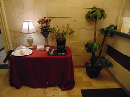 Una pequeña mesa con mantel rojo, un plato blanco con un pastel, un florero con flores, una cafetera y una planta en maceta.