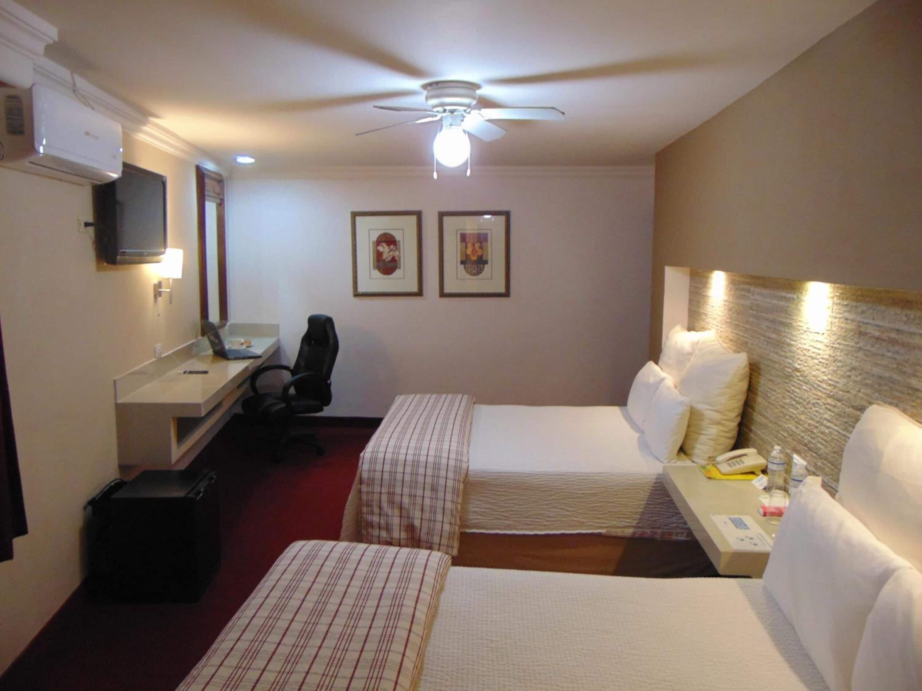 Habitación de hotel con dos camas, un escritorio con silla, ventilador de techo y dos cuadros en la pared.