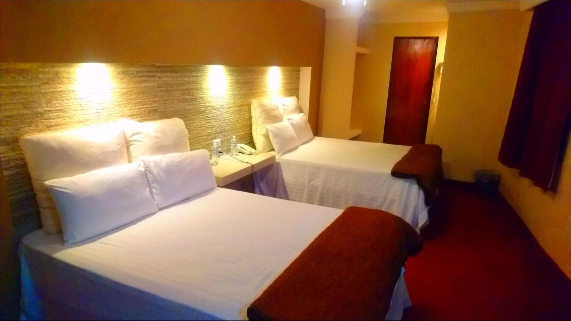 Hotellihuoneessa on kaksi sänkyä, joissa on valkoiset lakanat ja tyynyt. Huoneessa on pieni pöytä, jossa on puhelin ja vesipullo. Huoneen seinä on tehty tiilistä ja ovi on puuta.