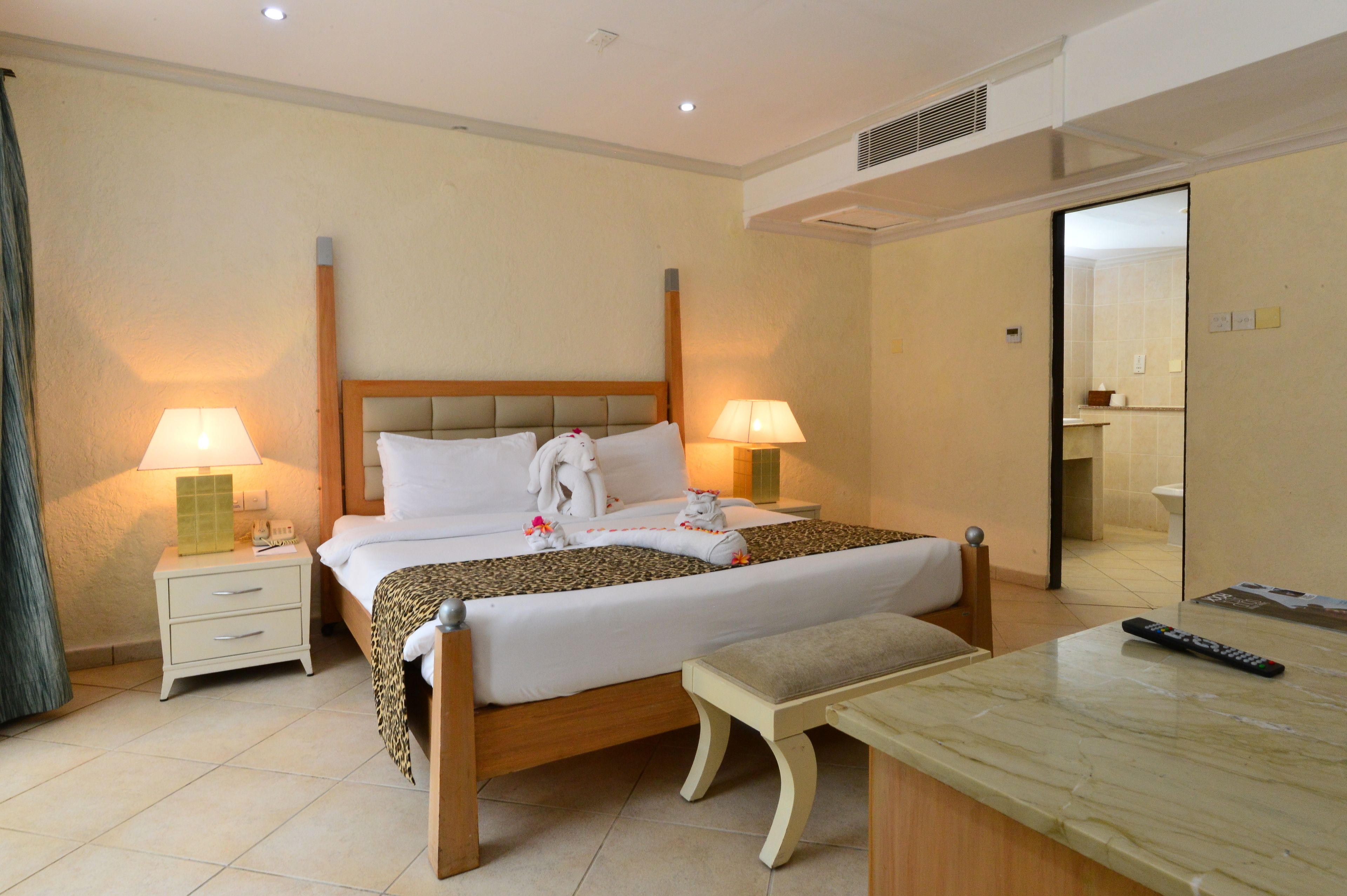 Habitación de hotel con una cama grande, dos mesitas de noche con lámparas, un banco y un control remoto sobre una mesa de mármol.