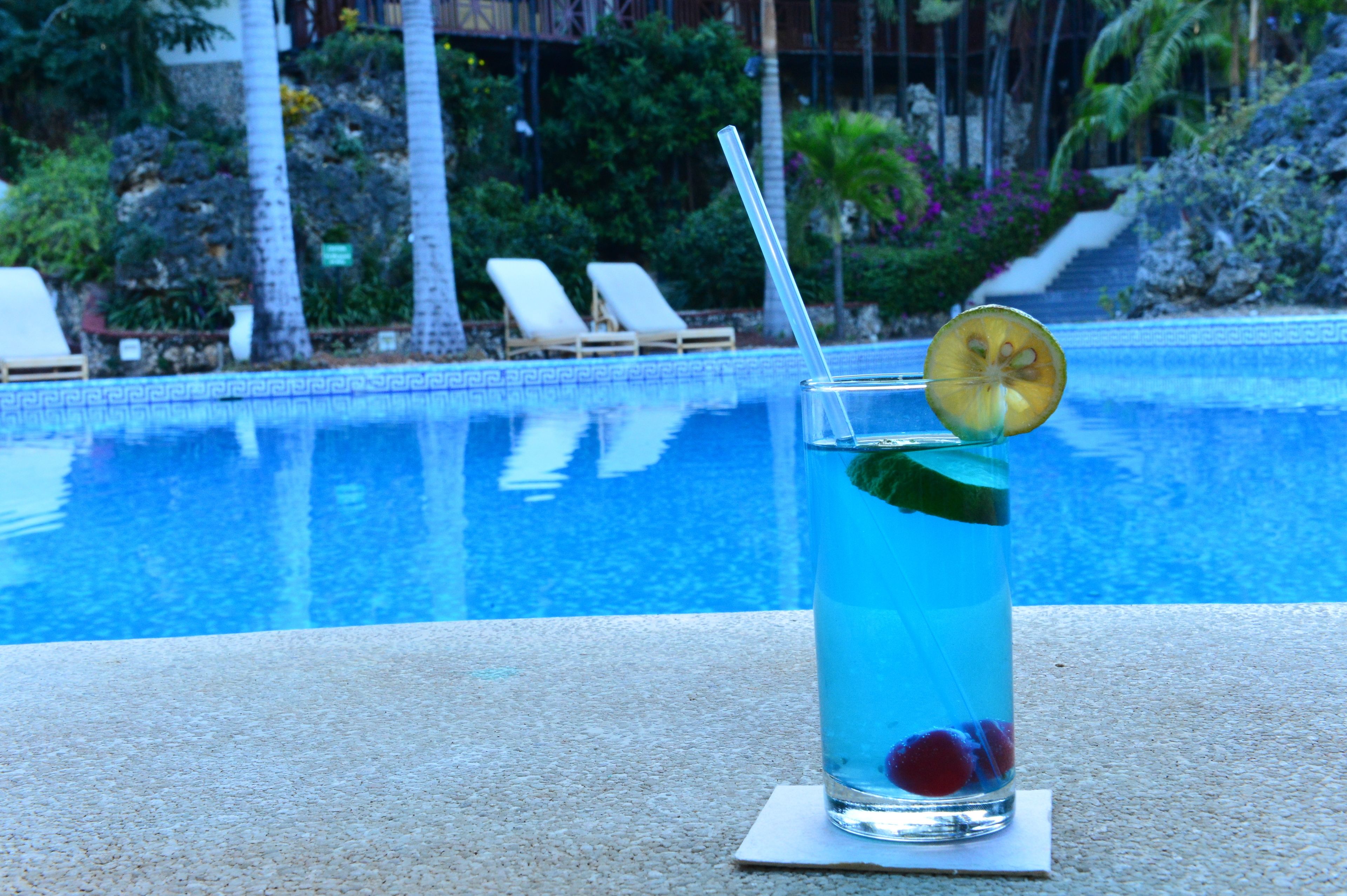 Un vaso de bebida azul con una rodaja de limón y hojas de menta sobre una mesa junto a la piscina.