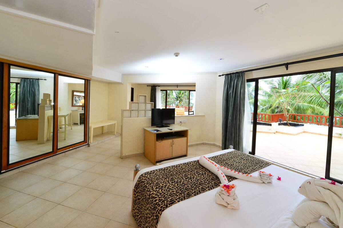 A hotel room with a large bed, a desk with a television, a mirror, and a balcony with a view of palm trees.