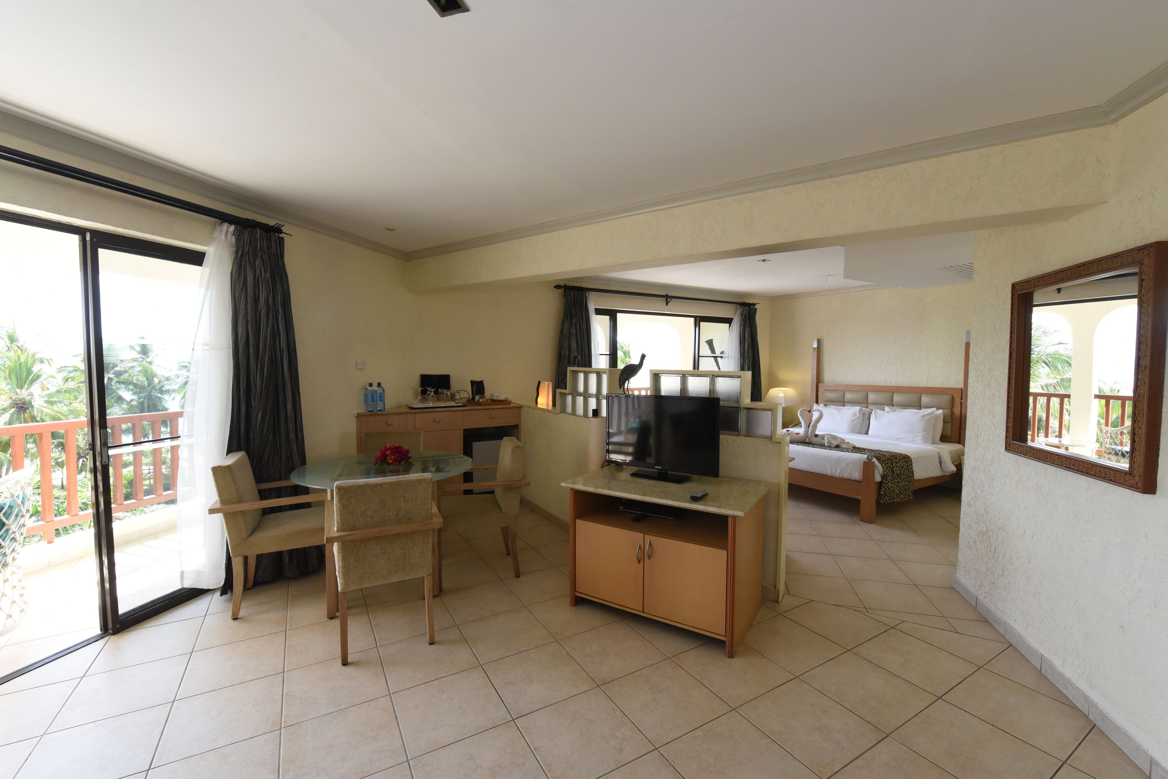 Habitación de hotel con balcón, zona de comedor, televisor, cama y escritorio.