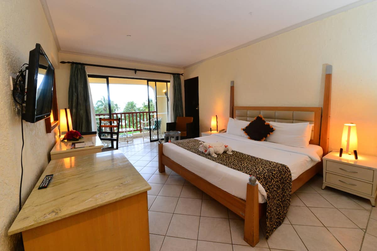 A hotel room with a bed, a desk with a TV, a chair, a nightstand, and a balcony with a view of palm trees.