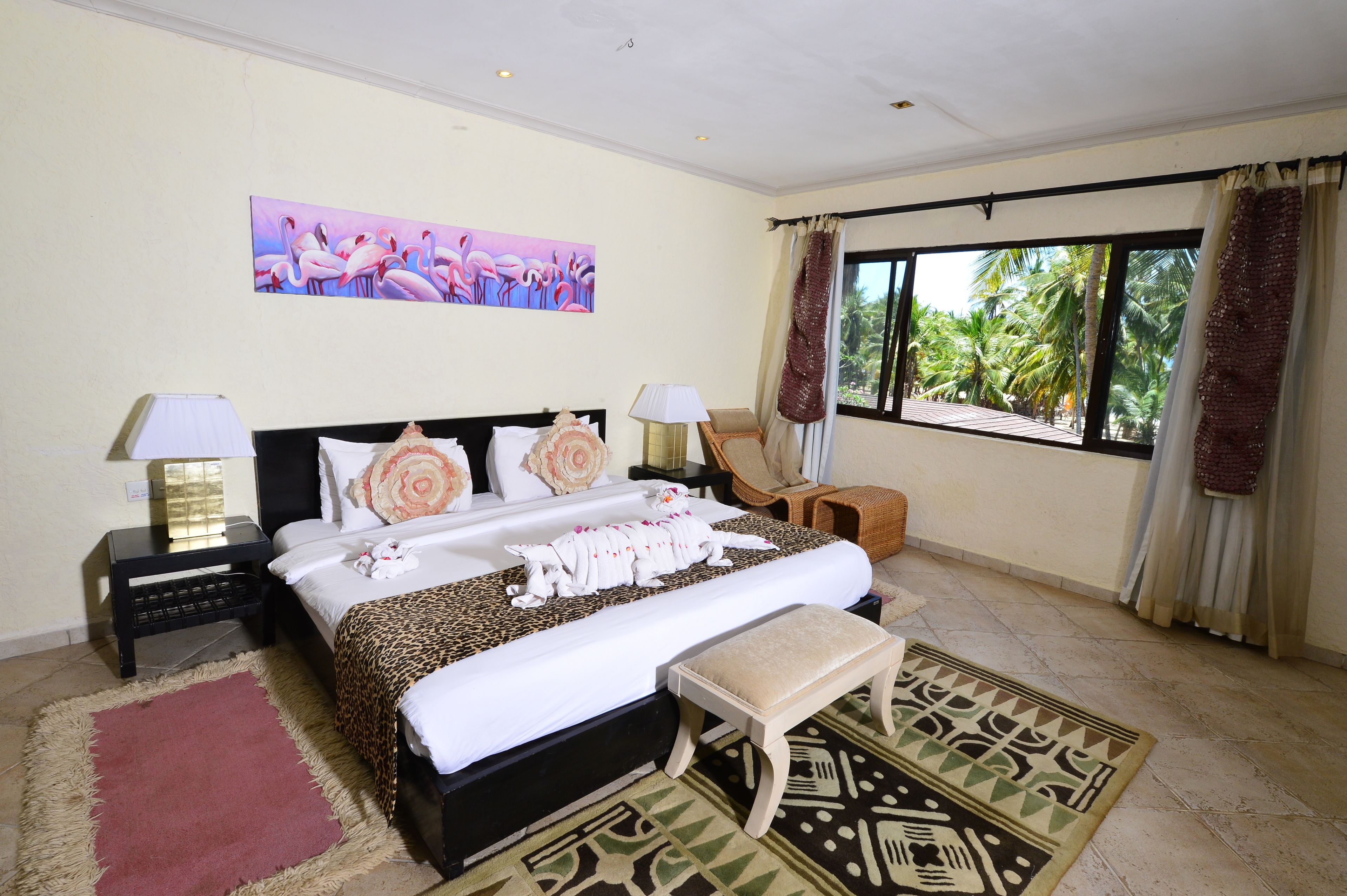 A hotel room with a large bed, two bedside tables, a nightstand, a bench, a patterned rug, a painting of flamingos, and a window with curtains.