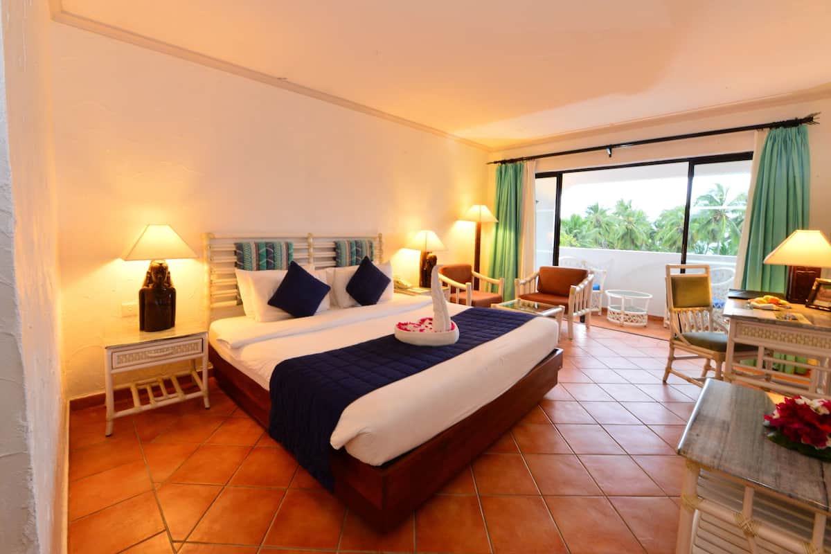 A hotel room with a large bed, two bedside tables, a desk, a chair, and a balcony with a view of palm trees.