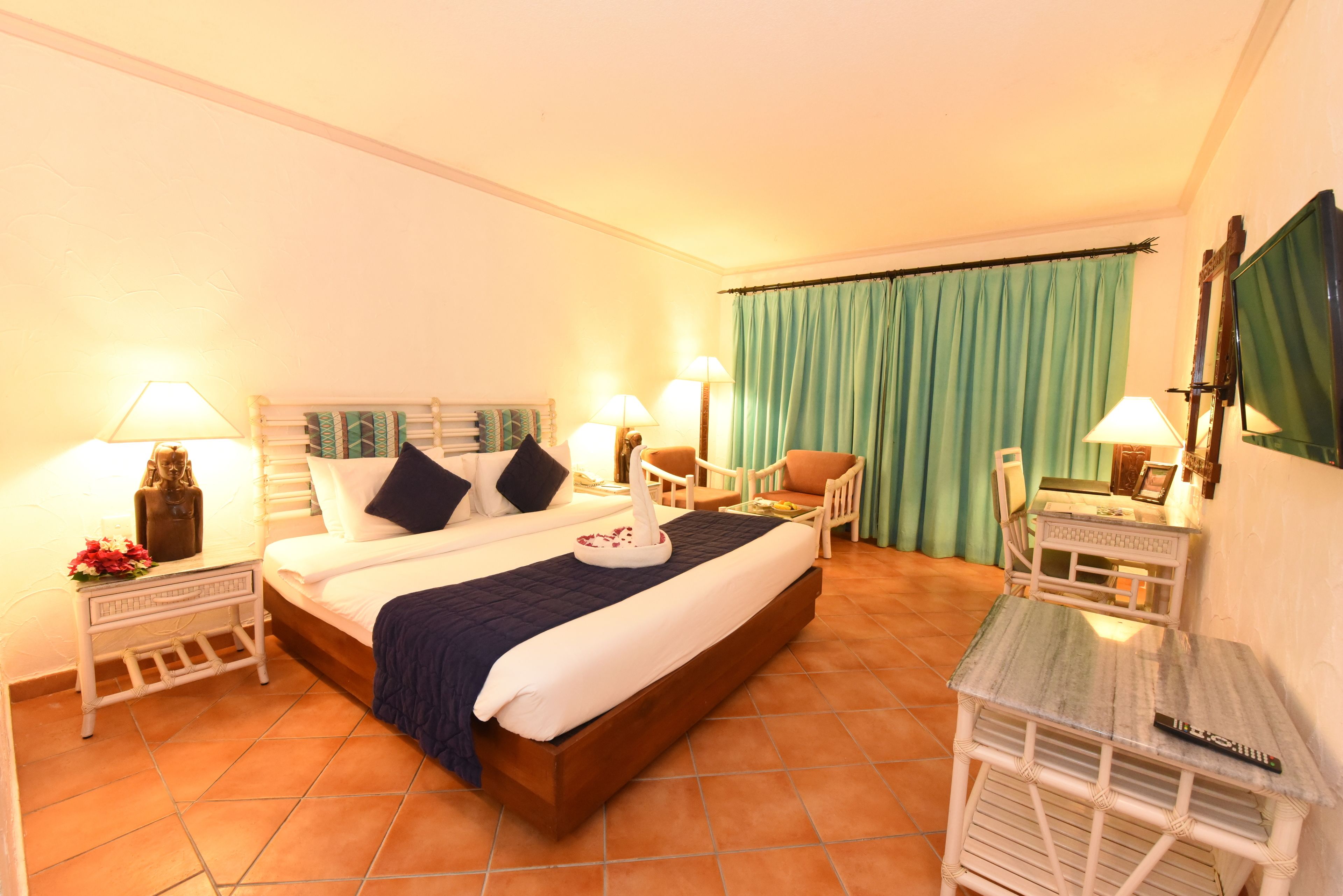 A hotel room with a large bed, two bedside tables with lamps, a chair, a small table, and a TV on the wall.