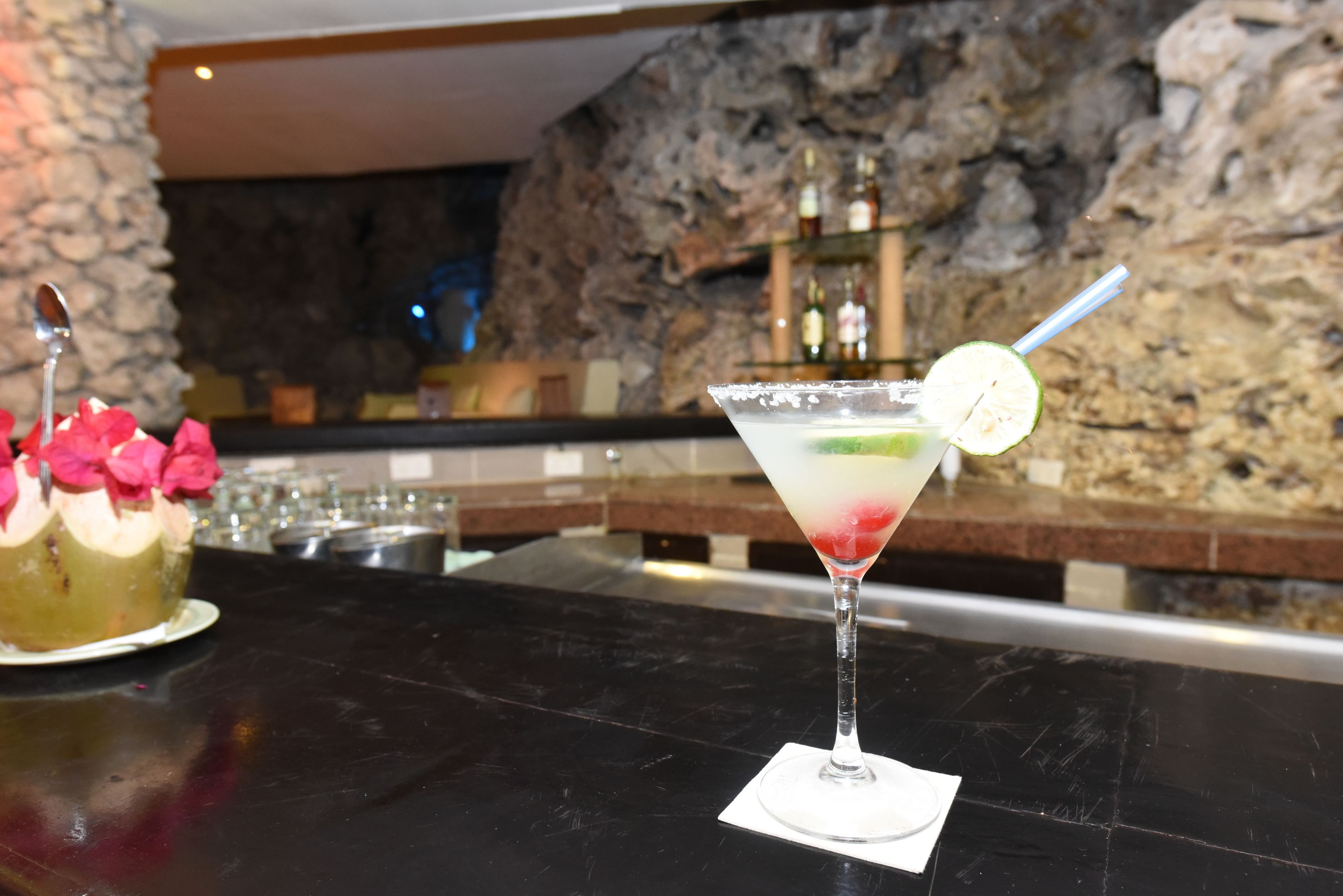Un coin bar avec un mur en pierre en arrière-plan, un verre à martini garni d’une tranche de citron vert et un gâteau décoré de fleurs roses, le tout posé sur un comptoir noir.
