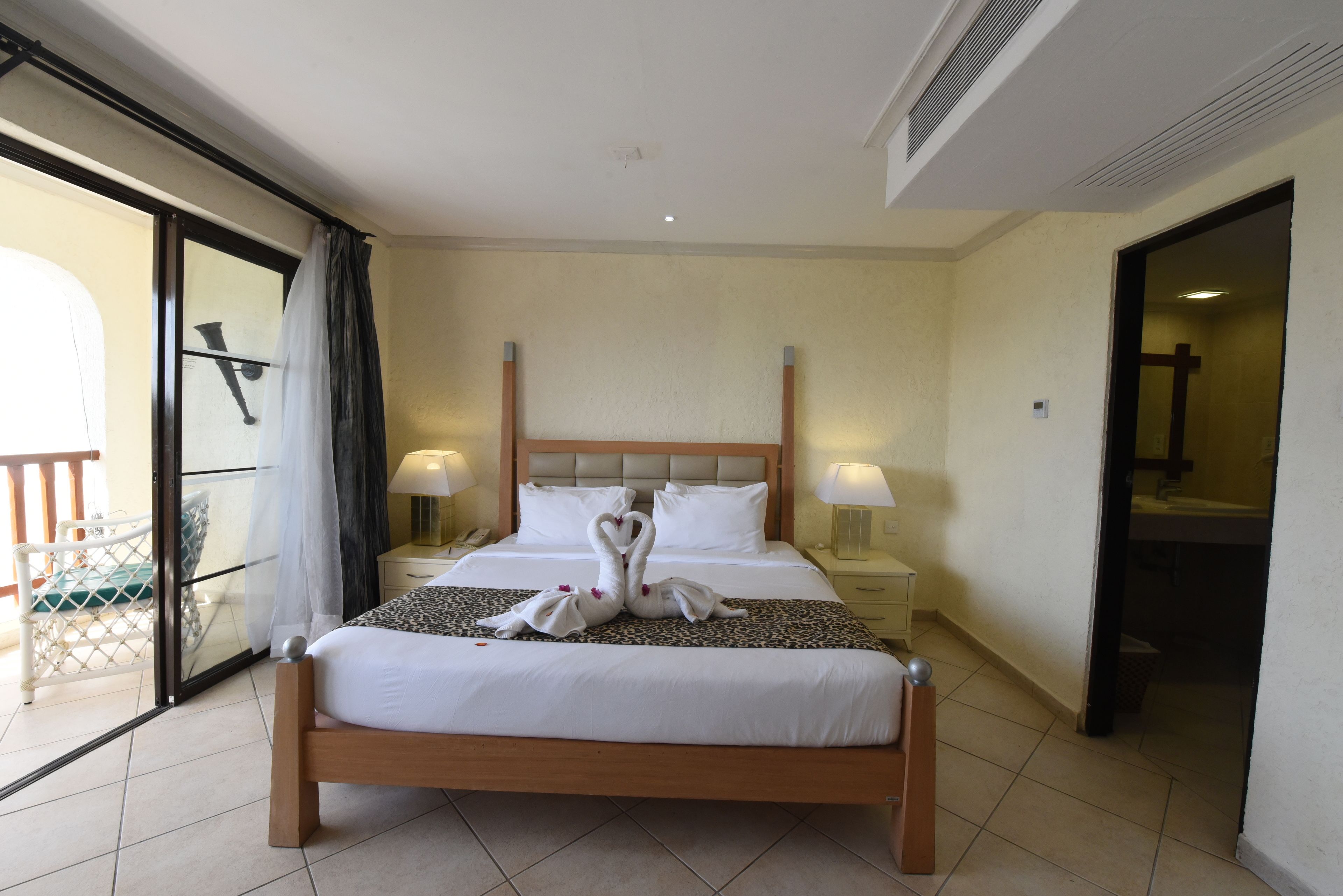 A hotel room with a large bed, two bedside lamps, a wooden headboard, and a view through a glass door to an outdoor area.