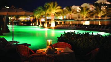A nighttime view of a pool area with lounge chairs and a table, surrounded by palm trees and illuminated by ambient lighting.