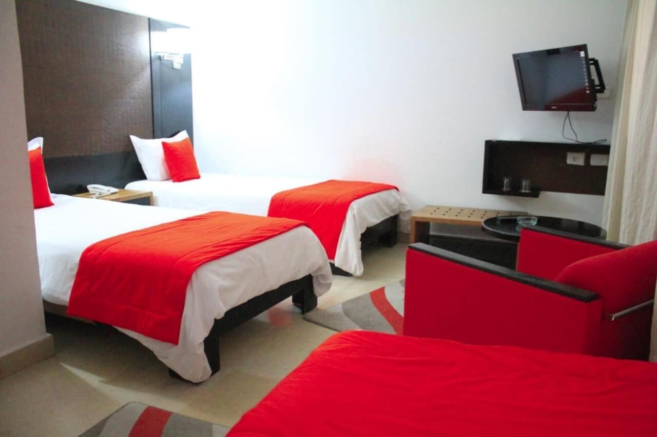 A hotel room with two beds, each with a red blanket, a small table, a TV mounted on the wall, and a red chair.