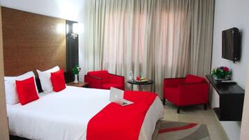 Une chambre d’hôtel avec un grand lit, des oreillers rouges et blancs, un fauteuil rouge, une petite table avec une plante et un téléviseur fixé au mur.