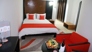 Camera d'albergo con un letto grande, un divano rosso, un tavolino con una bottiglia e bicchieri, e un vassoio di frutta.
