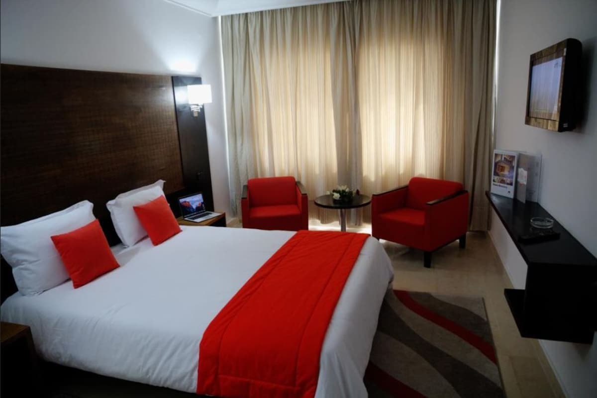 A hotel room with a large bed, two red chairs, a small table with a vase, and a TV on a stand.