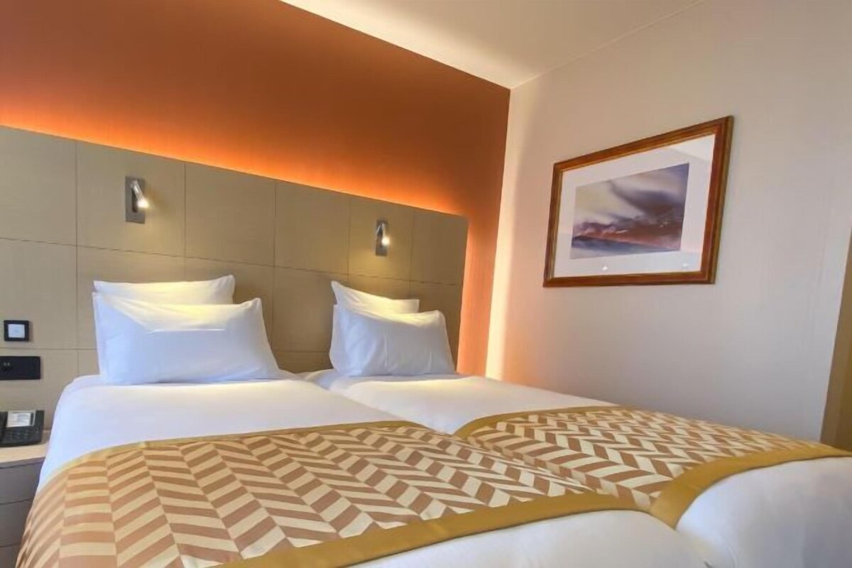 A hotel room with two beds, a headboard with integrated lighting, a framed picture on the wall, and a bedside table with a phone.