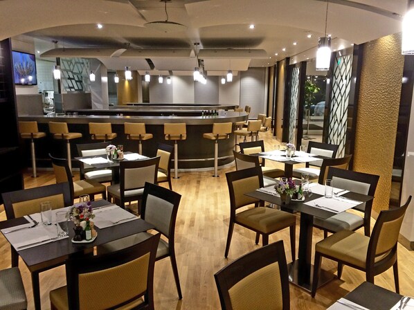 A modern restaurant with multiple dining tables and chairs, a bar area, and a kitchen in the background.