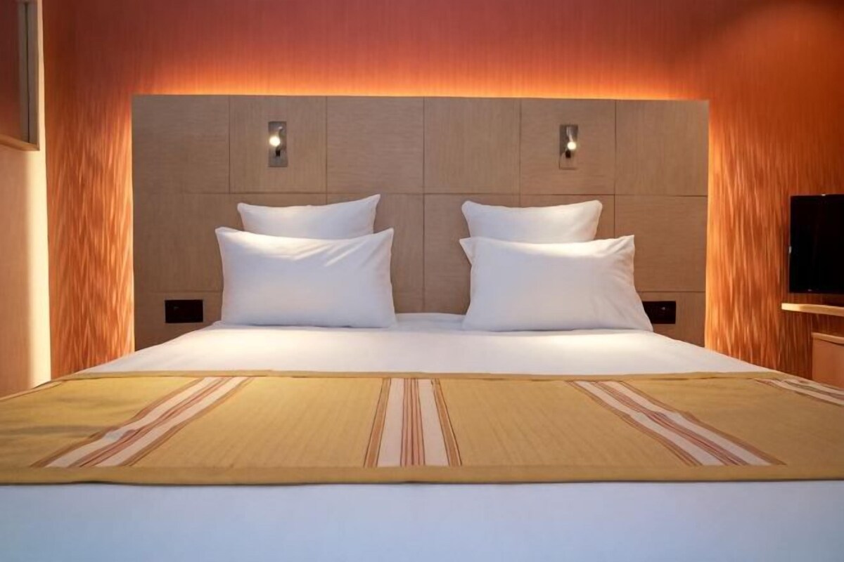 A large bed with white linens, a headboard with two wall-mounted lights, and a television on the right side.