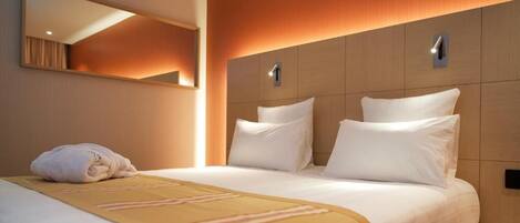 Habitación de hotel con una cama grande, cabecera con iluminación integrada, espejo y cama bien hecha con una toalla.