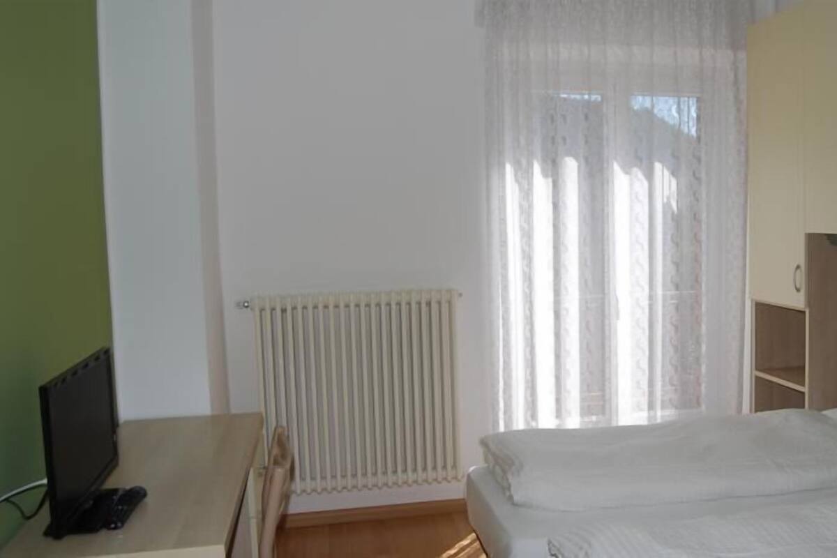 Un dormitorio con cama, un radiador, un escritorio con computadora y una ventana con cortinas.