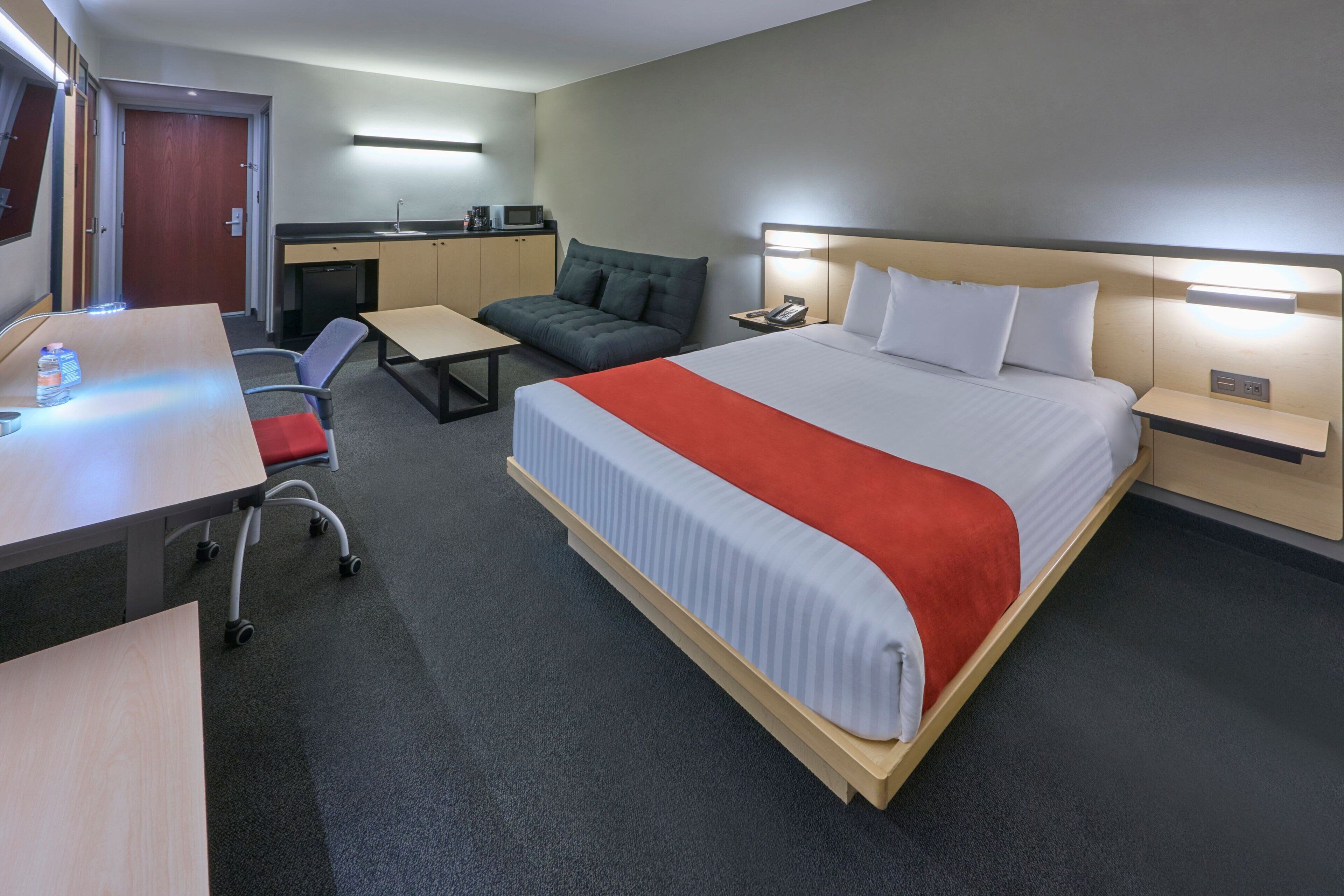 A hotel room with a bed, desk, chair, sofa, and kitchenette.