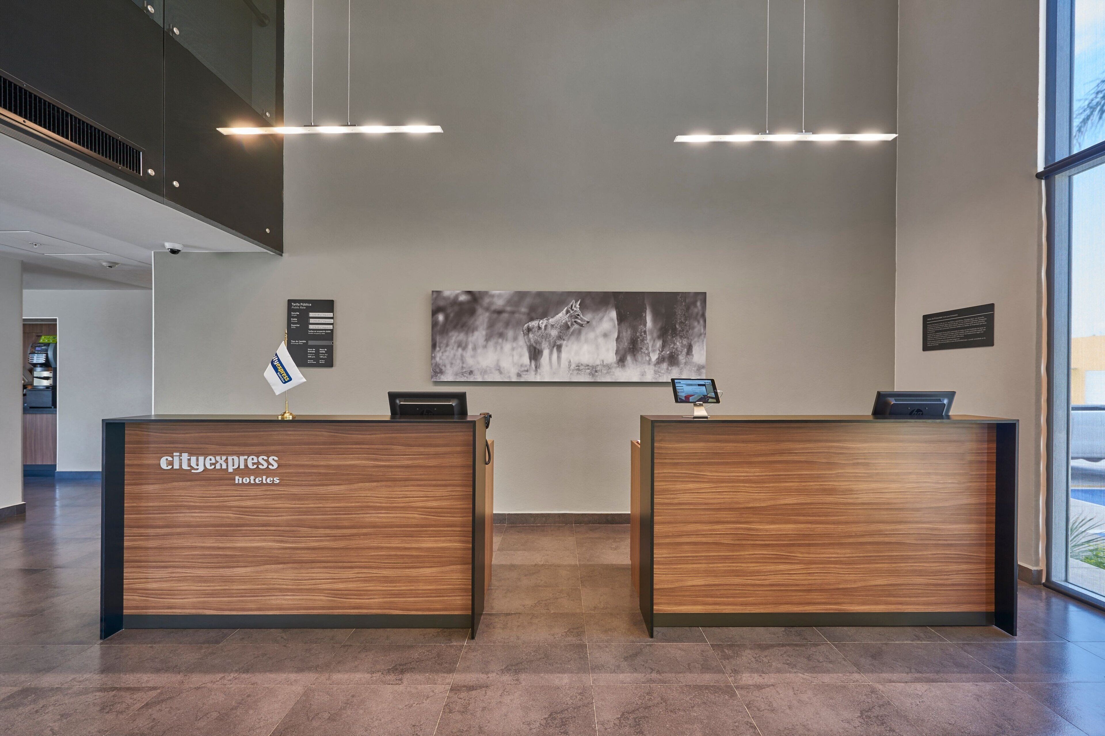 Un área de recepción con dos mostradores de madera identificados como "cityexpress hotels", una obra de arte blanco y negro en la pared y una pantalla digital en uno de los mostradores.
