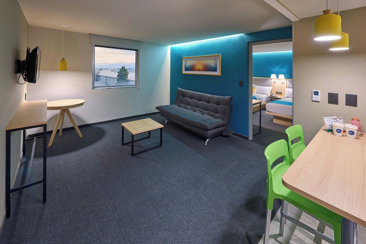 Une chambre d’hôtel moderne équipée d’un canapé, d’une petite table, d’une table à manger avec des chaises, d’un téléviseur fixé au mur et d’une fenêtre donnant sur un paysage agréable.