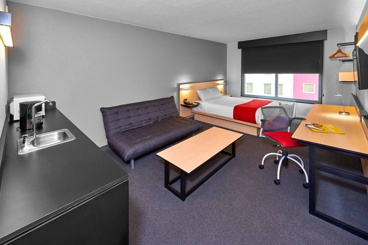 Une chambre d’hôtel de taille réduite, comprenant un lit, un canapé, un bureau, une chaise, une petite table, un lavabo et un téléviseur.