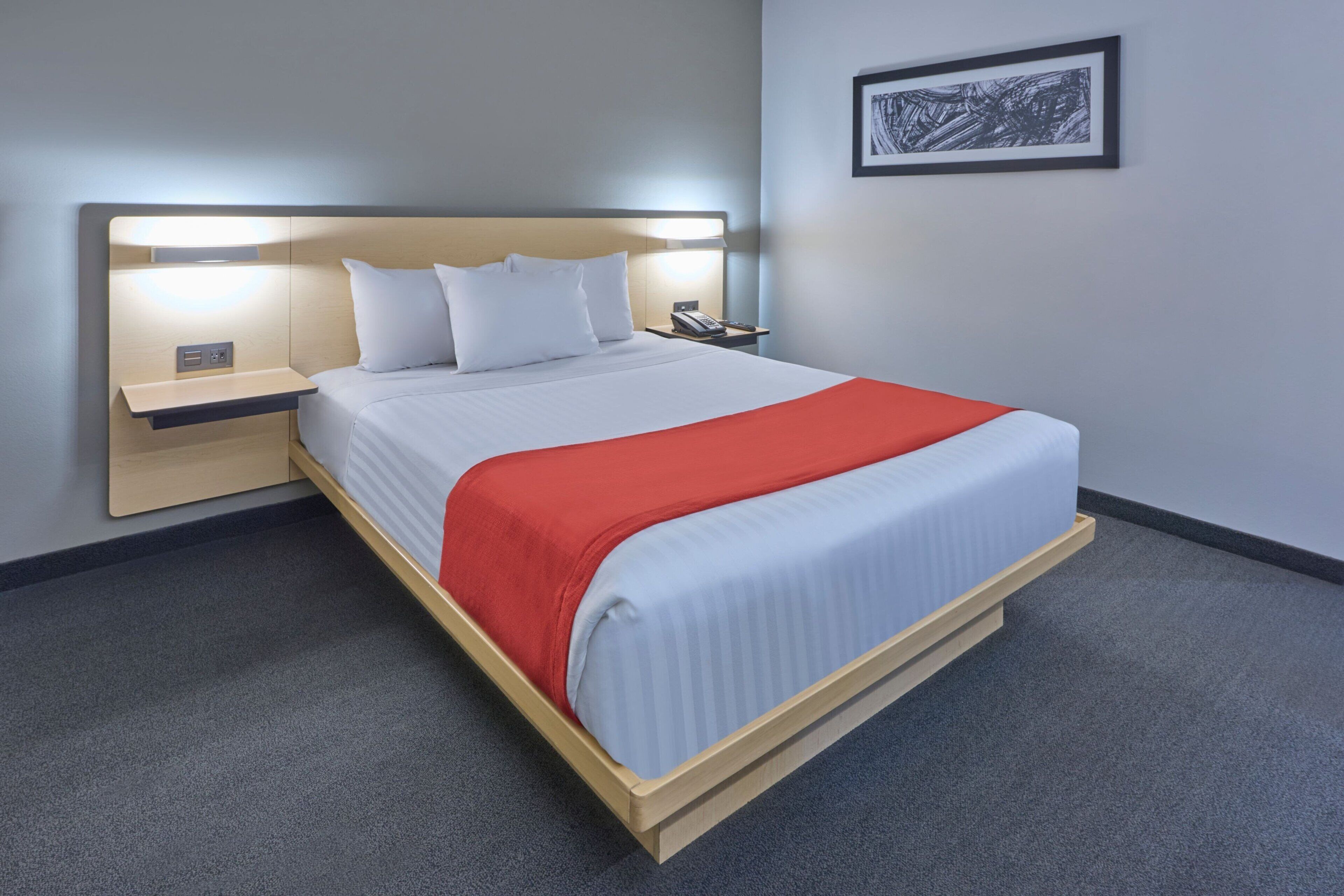 Una habitación de hotel moderna con una cama grande, una mesita de noche con una lámpara y un cuadro enmarcado en la pared.