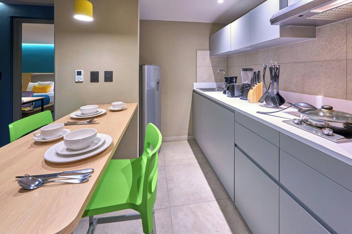 Une chambre d’hôtel compacte dotée d’un coin repas avec une table en bois, dressée avec de l’assiette blanche, des bols et des couverts. Une chaise verte est placée à la table. La chambre comprend un coin cuisine équipé d’appareils électroménagers modernes et un plan de travail sur lequel sont disposés divers ustensiles de cuisine.