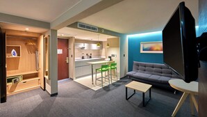 A modern living room with a grey sofa, a coffee table, and a TV. There is a kitchen area with a dining table and chairs. - City Express by Marriott Toluca (Toluca)