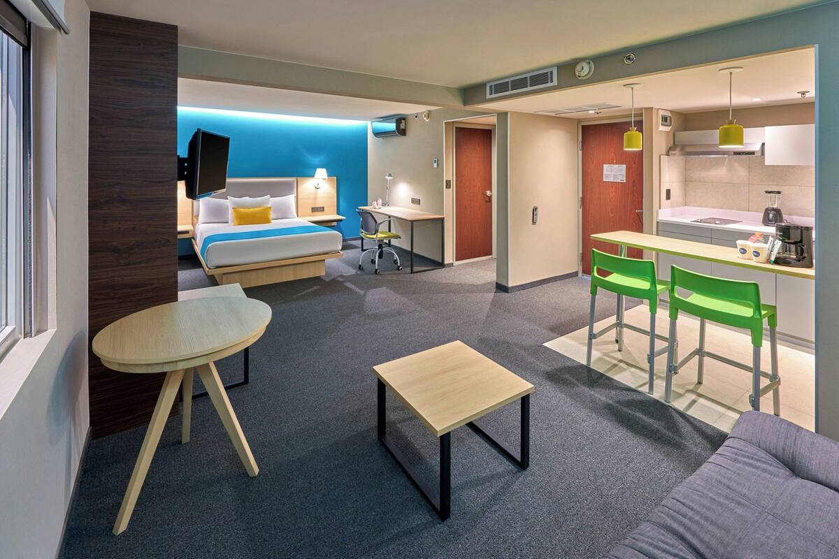 Une chambre d’hôtel moderne équipée d’un lit, d’un bureau, d’une chaise, d’une petite table et d’une kitchenette.