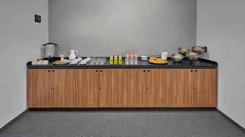 A buffet setup with various food items, beverages, and utensils on a counter against a grey wall.