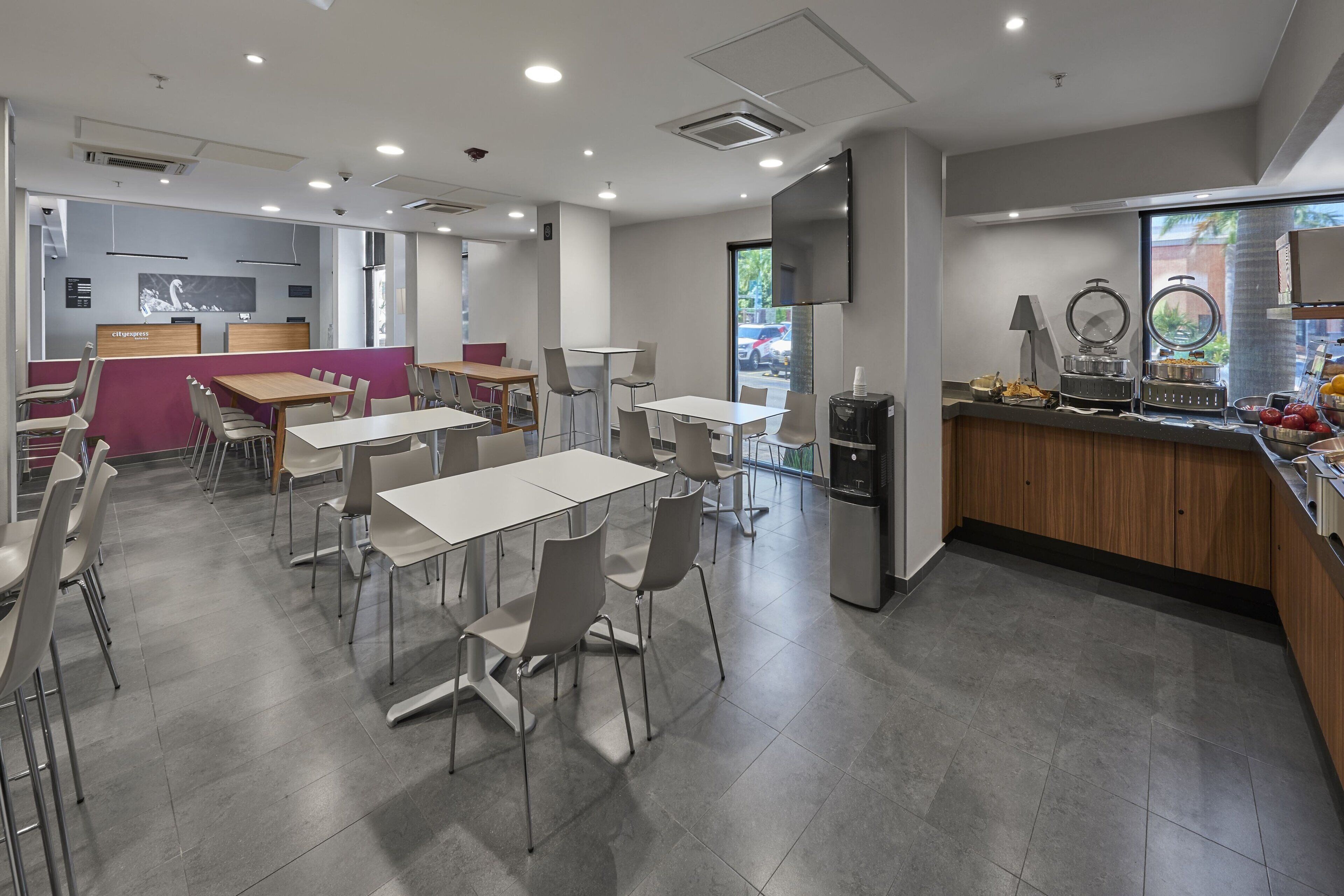 Un comedor moderno con mesas y sillas, una encimera de cocina con alimentos y un amplio ventanal con vistas al exterior.