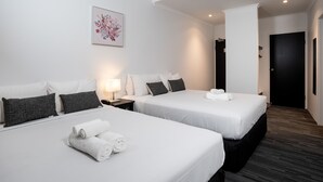 Quarto de hotel com duas camas, roupa de cama branca, almofadas cinza escuro, uma pequena mesa de cabeceira com abajur e uma pintura floral emoldurada na parede.
