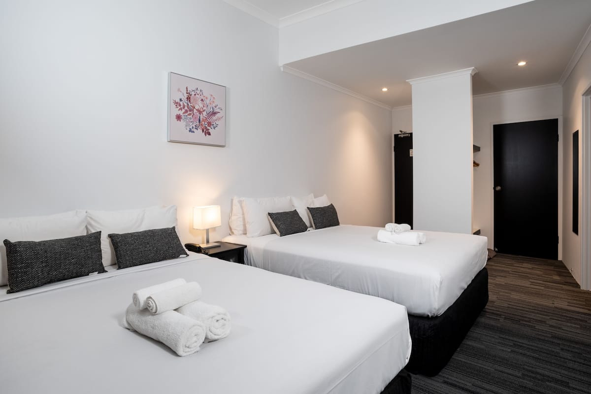 A hotel room with two beds, white linens, dark gray pillows, a small bedside table with a lamp, and a framed floral artwork on the wall.