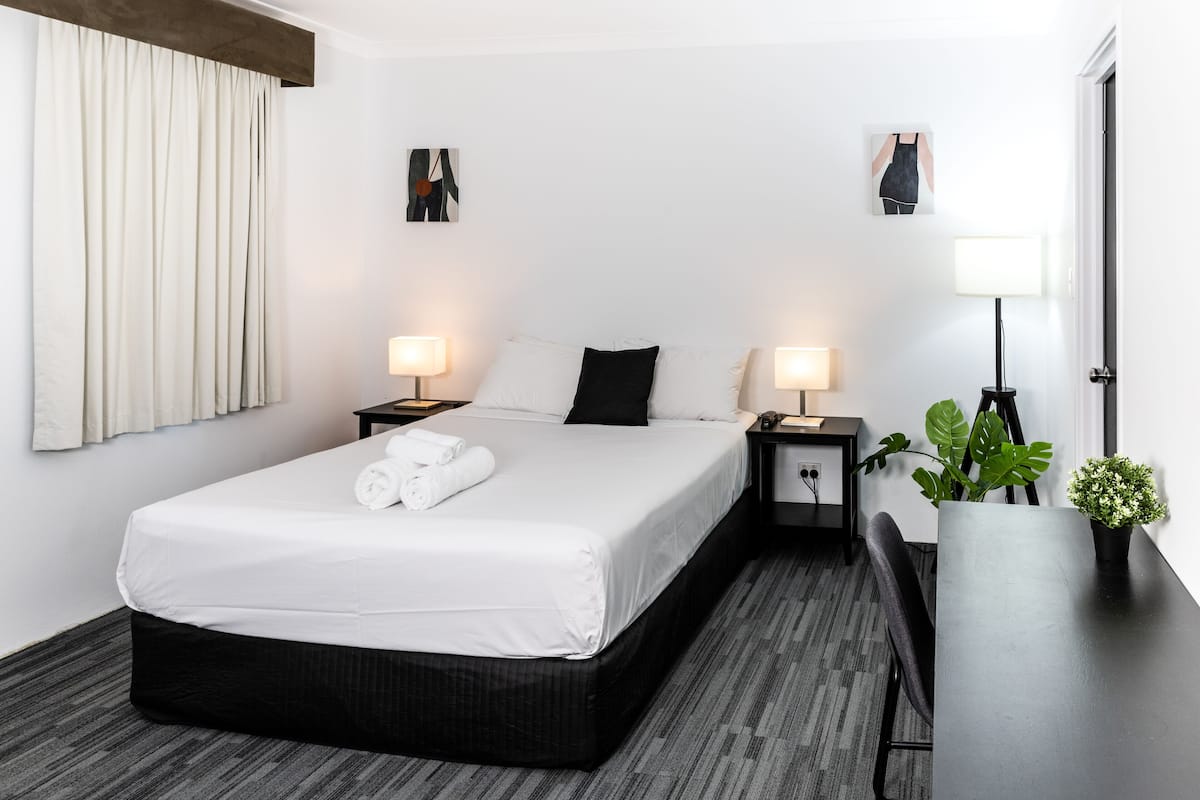 A hotel room with a bed, bedside tables, lamps, a desk, and a potted plant.