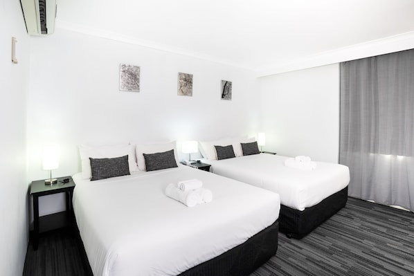 A hotel room with two beds, white linens, dark headboards, bedside tables with lamps, and a grey curtain. - Joondalup City Hotel (Joondalup)