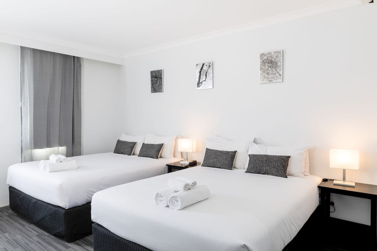 A hotel room with two beds, each with white linens and rolled towels, a nightstand with a lamp, and three framed artworks on the wall.