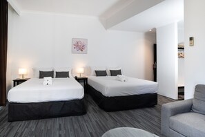 A hotel room with two beds, a gray armchair, a small round table, and a wall-mounted artwork. - Joondalup City Hotel (Joondalup)