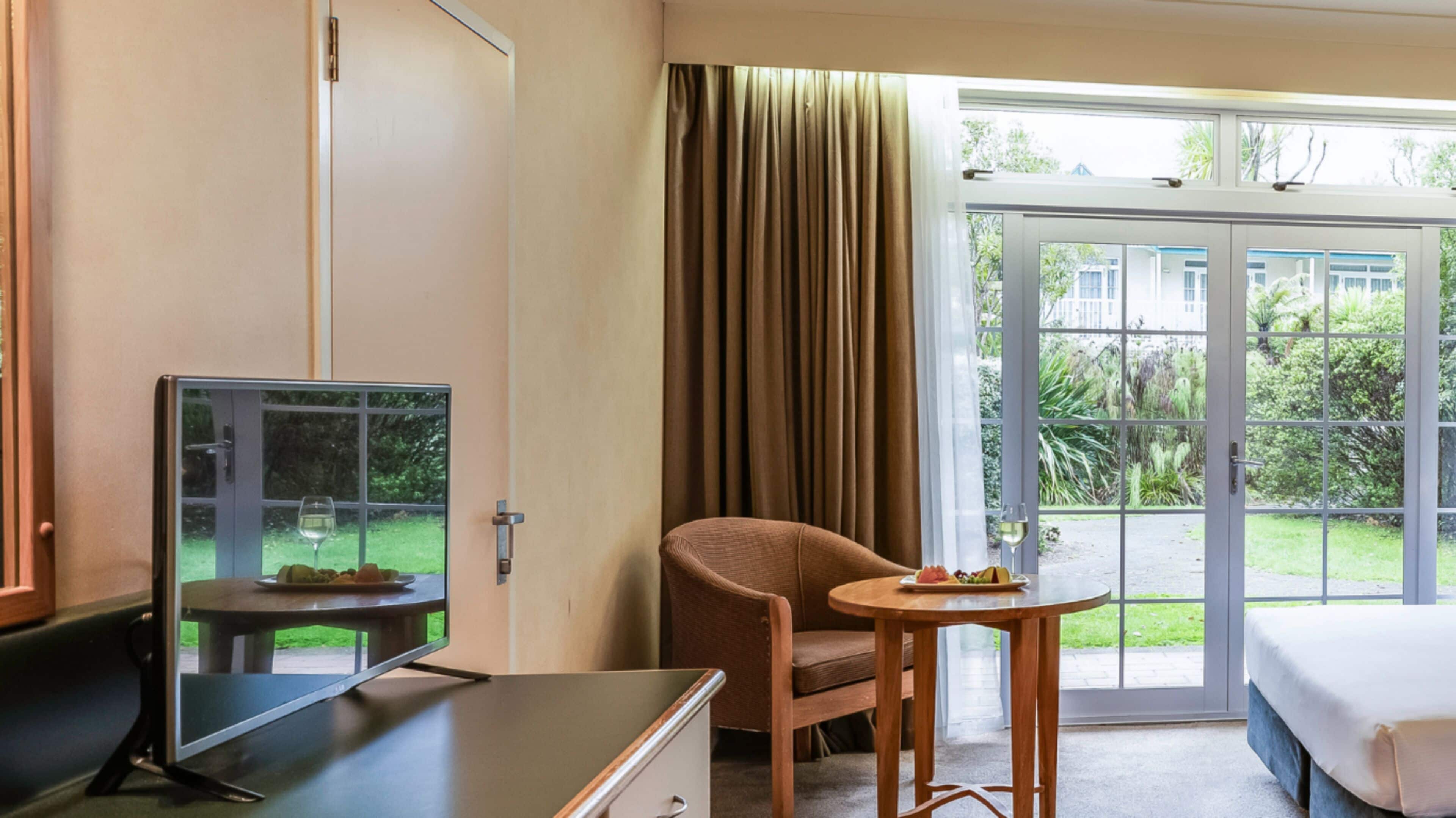 Ein Hotelzimmer mit Bett, Schreibtisch mit Computer, Stuhl, kleinem Tisch mit einem Weinglas und Blick auf einen Garten durch große Fenster.