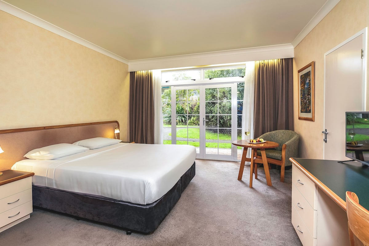 Een hotelkamer met een bed, nachtkastje, lamp, bureau, stoel en uitzicht door een glazen deur naar buiten.