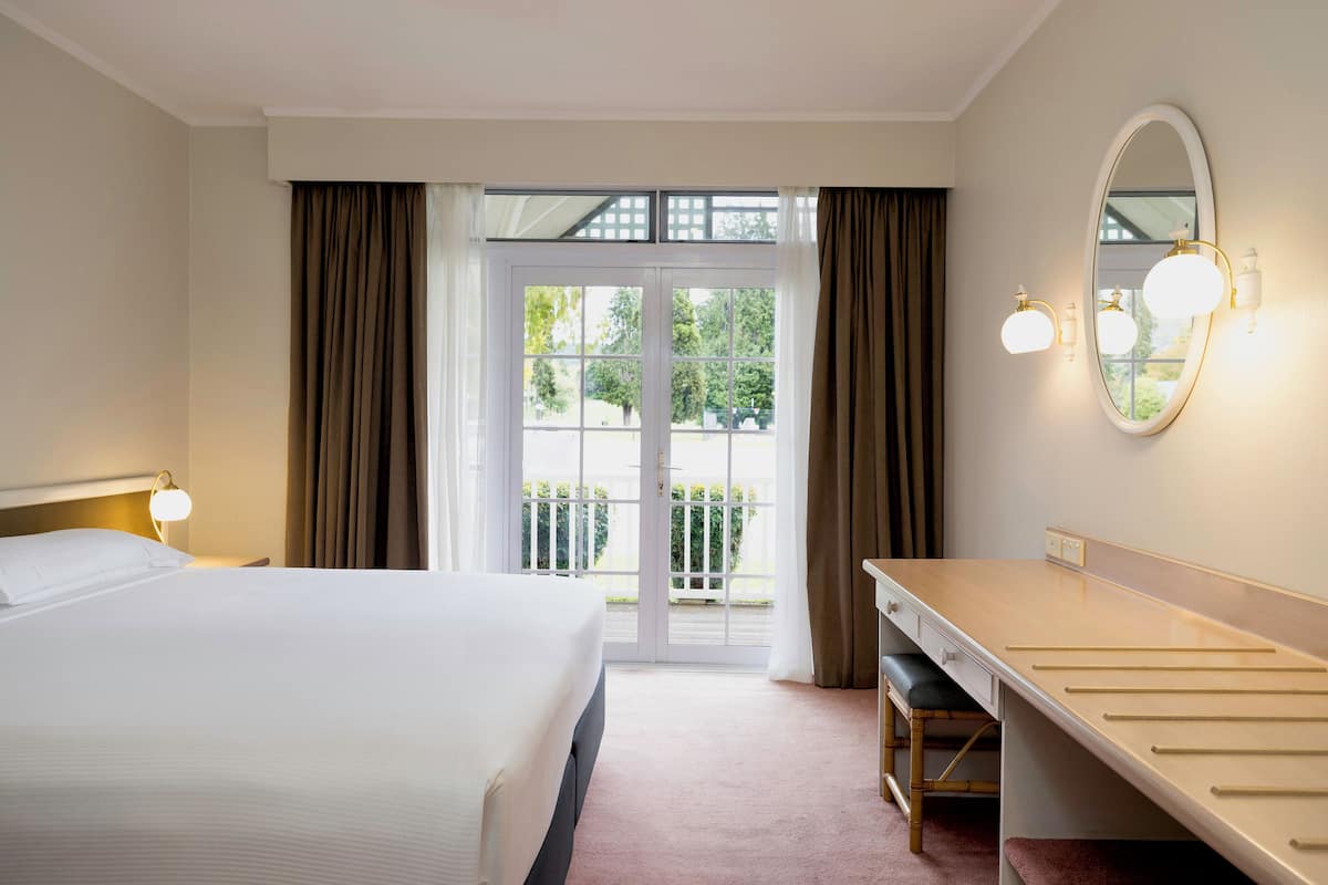 Een hotelkamer met een bed, een bureau, een spiegel en uitzicht op het groen door een glazen deur.