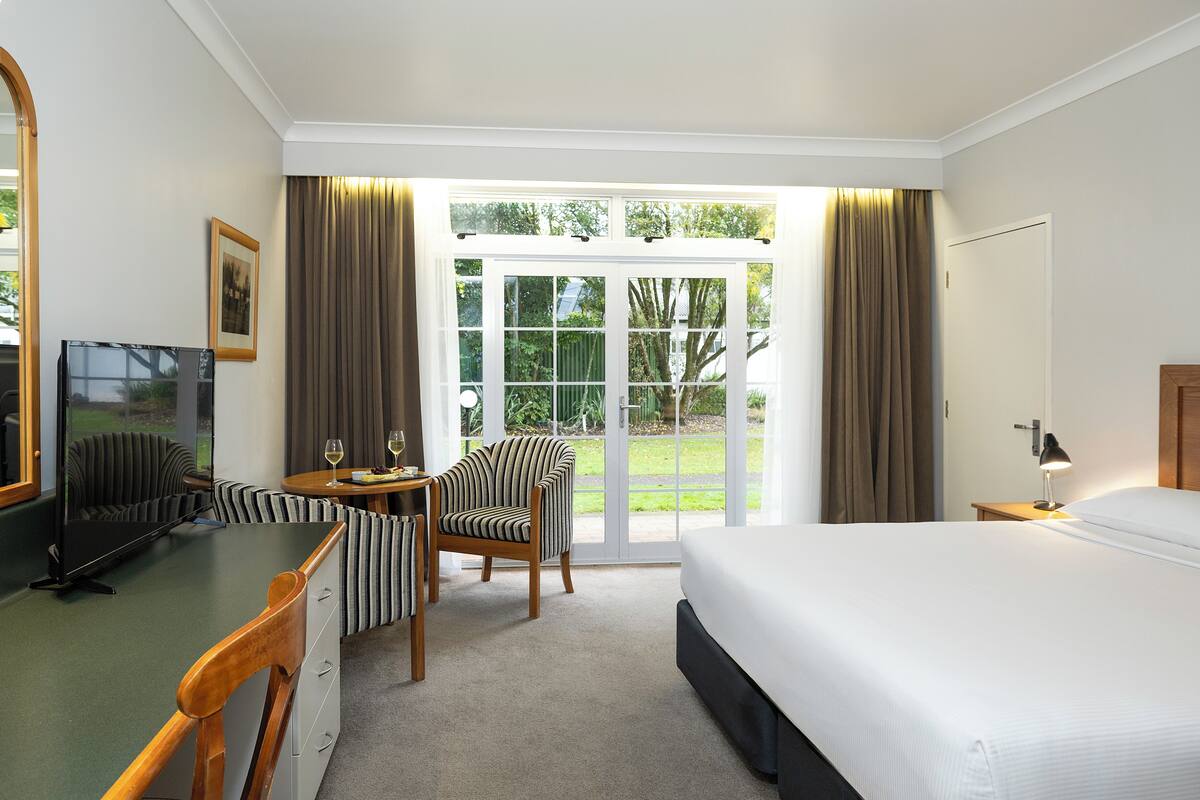 Een hotelkamer met een bed, een bureau met een stoel, een televisie en uitzicht op het landschap door grote glazen deuren.